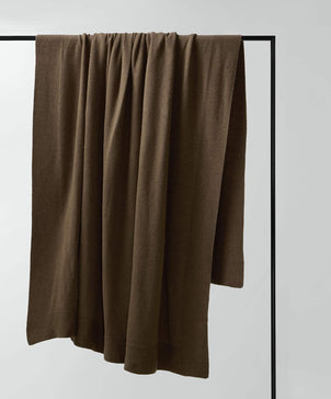 A 100% NZ Merino Wool Throw in Truffle is draped over a sleek black metal frame against a white background, highlighting its refined New Zealand craftsmanship.