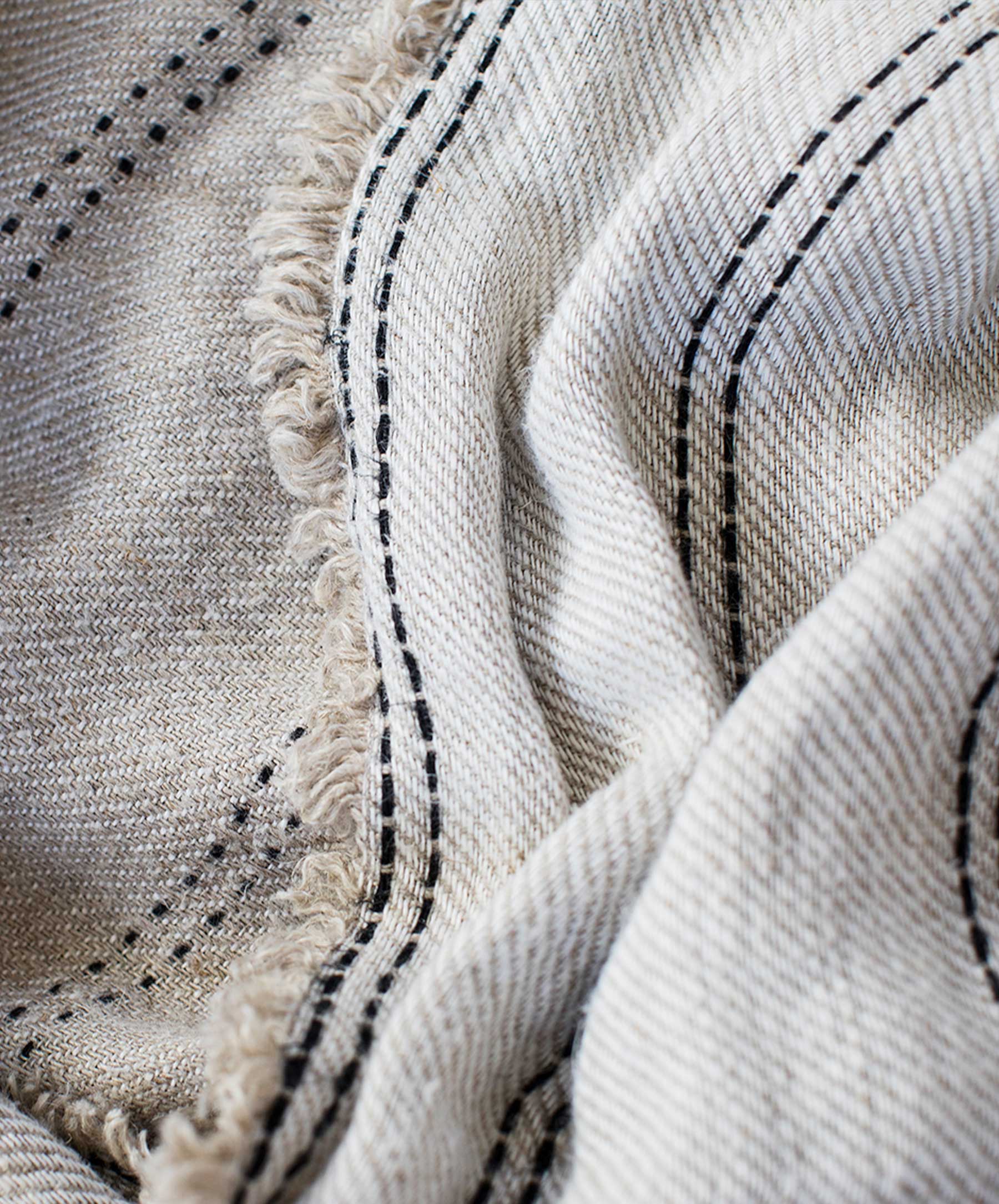 Close-up of "A pairing for quiet retreat - Mira & Steel Blue": textured beige fabric of natural fibres, black stitched lines, frayed edge; the weave and soft folds evoke calm and comfort.