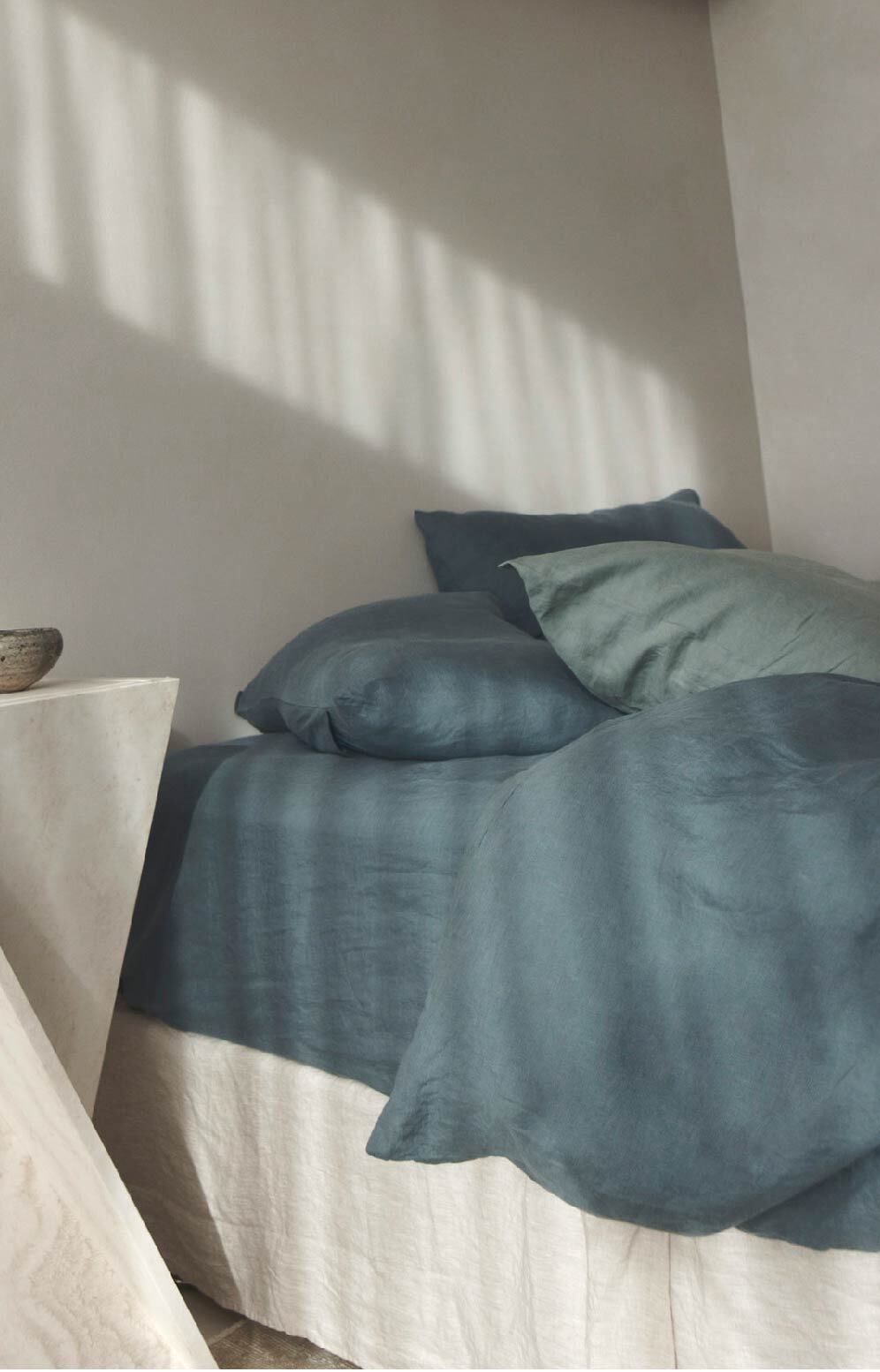 A neatly made bed with blue-gray linen bedding and pillows, set against a white wall with soft shadows from window blinds. A white nightstand is partially visible on the left, holding a small bowl.