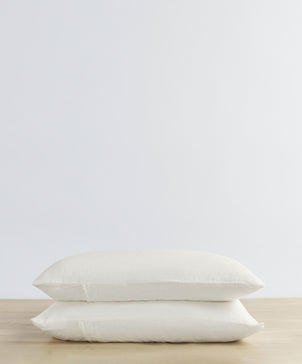 The Set of 2 Linen Pillowcases in Ivory are stacked neatly on a light wooden surface, creating a vintage look against a plain white background.