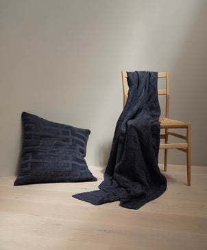 Dark blue textured pillow and blanket on a wooden chair against a plain wall.