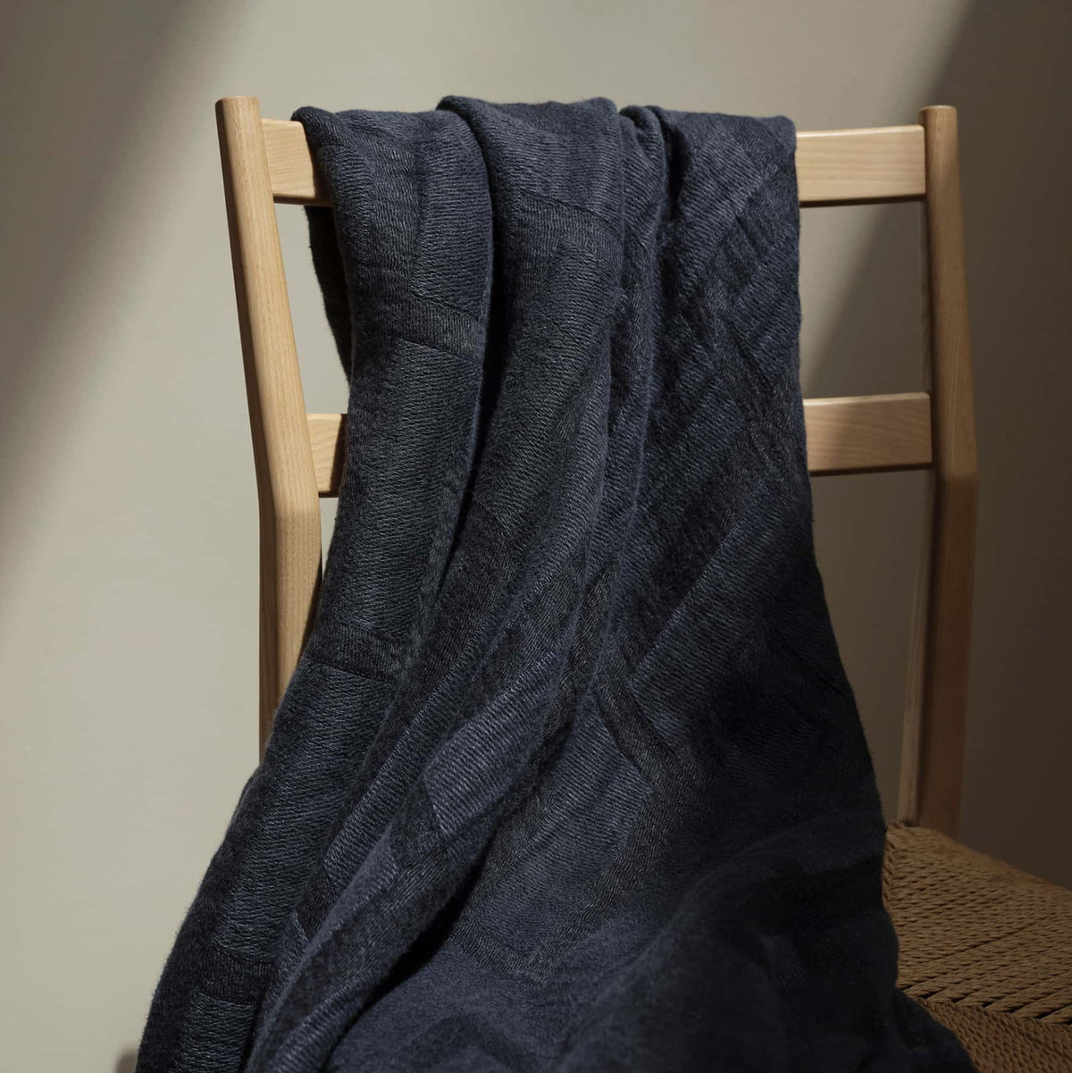 A dark textured blanket draped over the back of a light wooden chair with a woven seat, under soft indoor lighting.