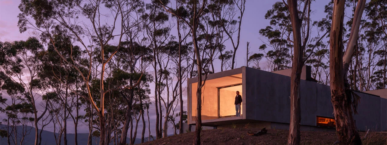 A modern concrete cabin with a large open front wall, glowing warm light inside, set among tall trees at dusk under a purple sky. A person stands in the doorway.