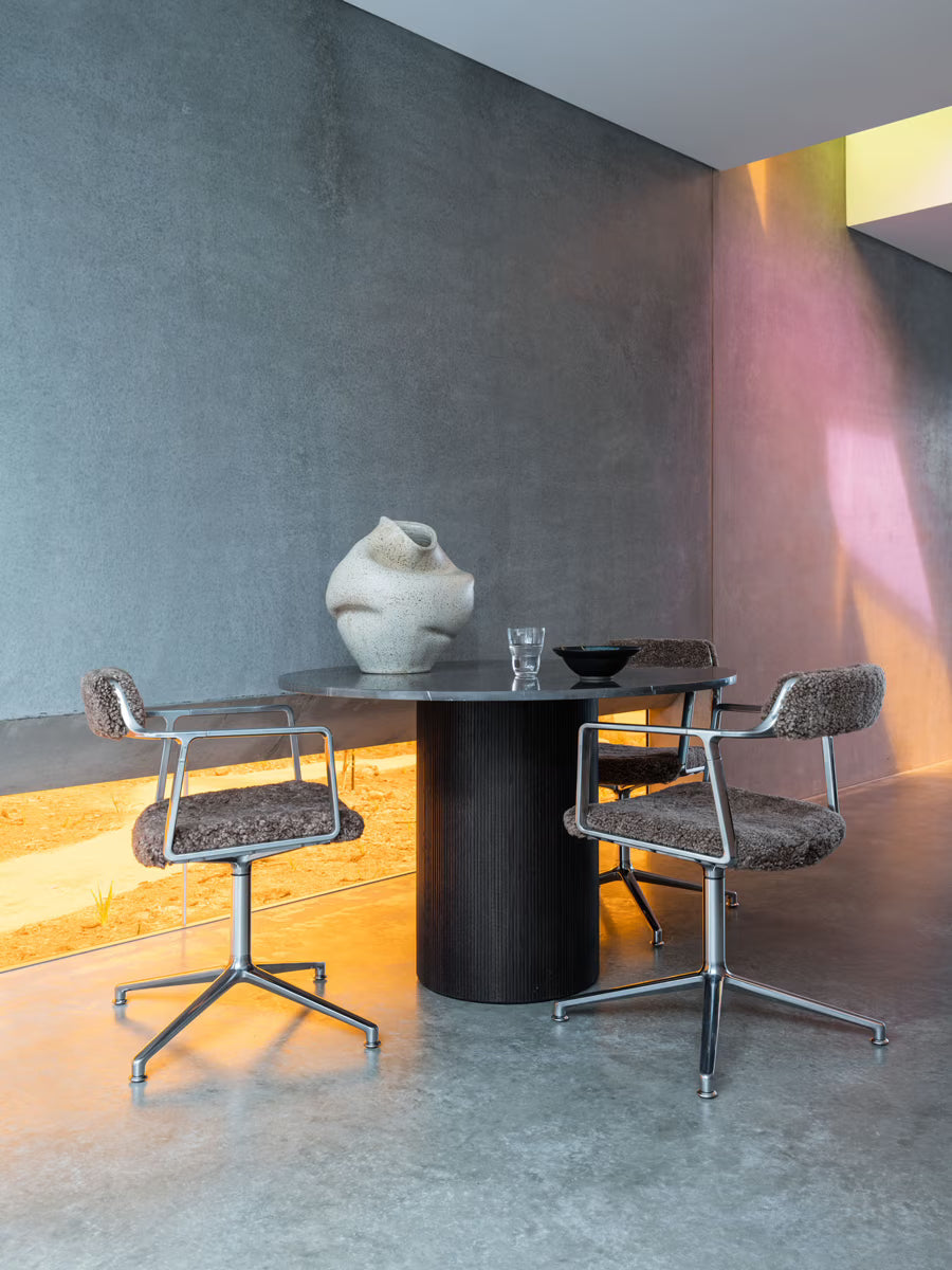 Modern dining set with two textured chairs and a round, dark table holding a sculptural vase, glass, and bowl. The background features a concrete wall and warm, yellow lighting at the base.