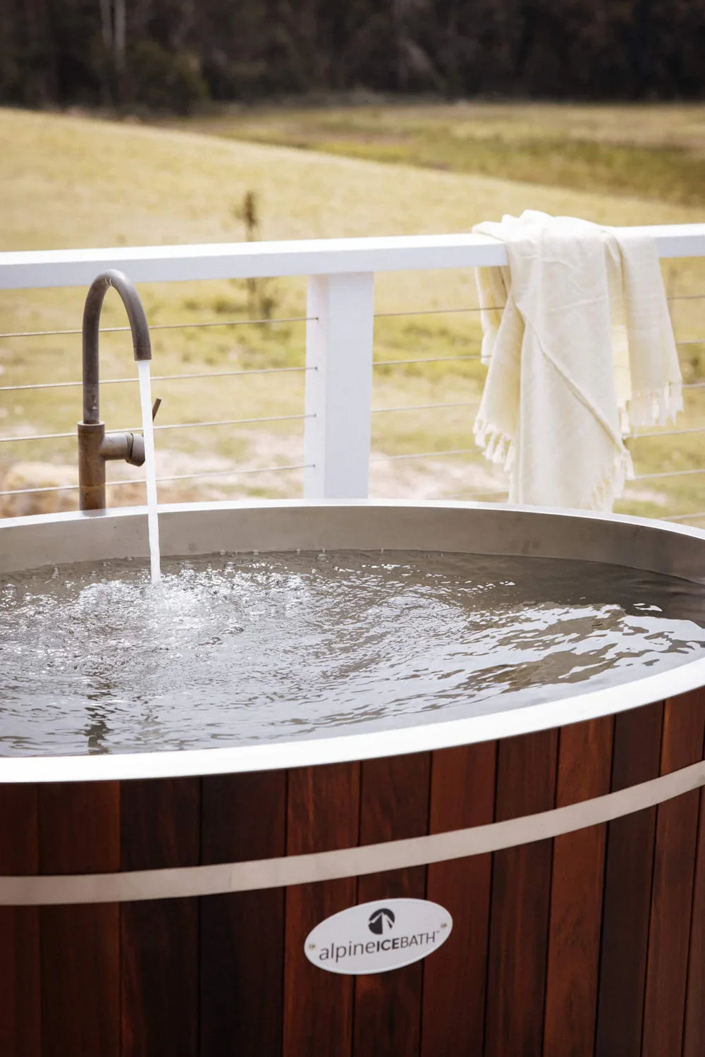An outdoor round hot tub with dark wood paneling and a chrome faucet, shown with water running in. A white towel hangs over a white railing in the background, with a grassy field and trees beyond.