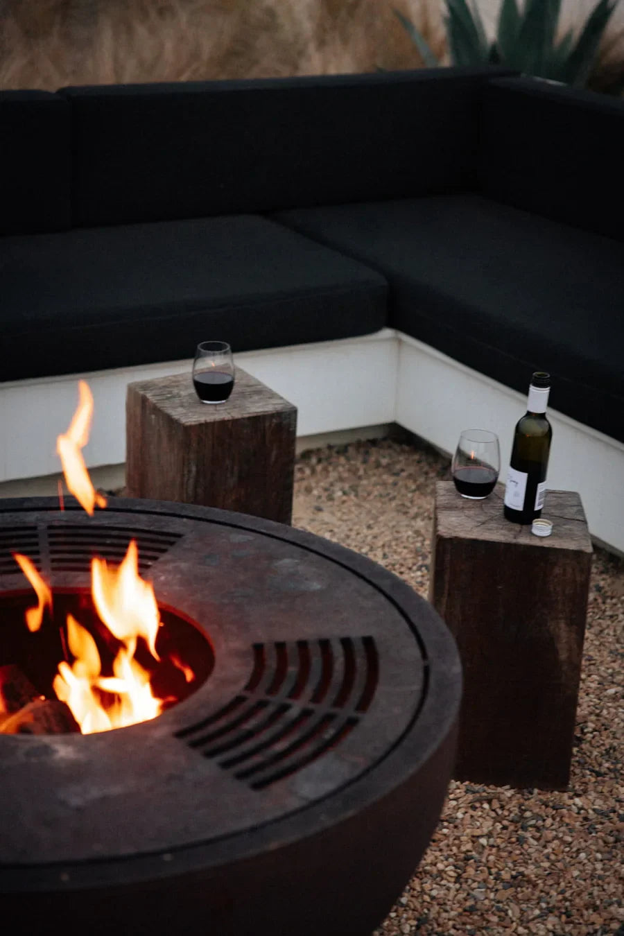 A round dark metal fire pit with visible flames in the foreground, set in a patio environment with a black sectional sofa, wooden side tables, and a wine bottle on a gravel surface.