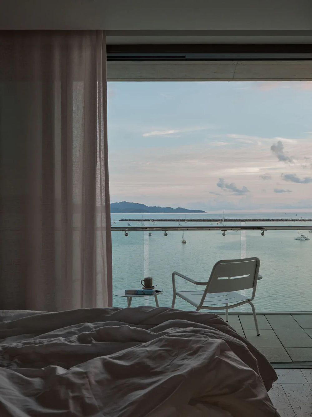 Room with a bed and a chair overlooking a body of water with mountains in the distance.