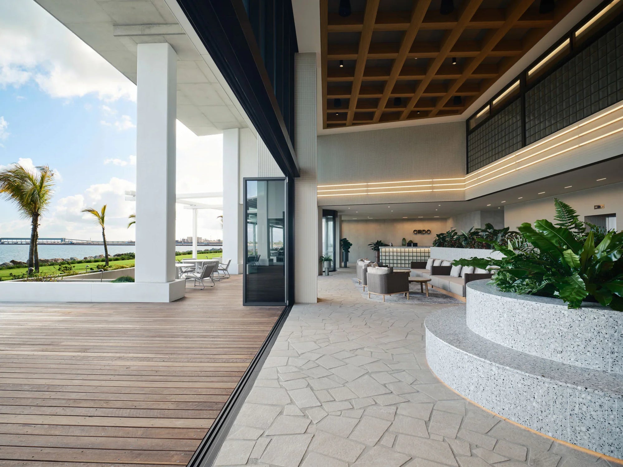 Modern indoor-outdoor area with wooden deck, glass walls, and greenery.