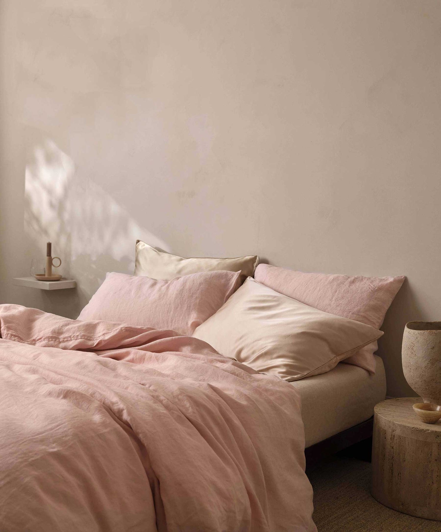 A cozy minimal bedroom features a bed with blush and beige bedding, including the Silk Linen Flip Pillowcase - Blush. Pillows rest against a cream wall as soft light enters. A vase and candle sit on simple bedside tables.