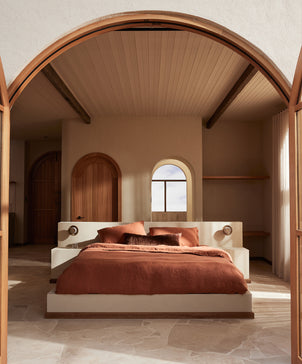 A minimalist bedroom with a modern, earthy design features a low white platform bed accented by the Talik Velvet Lumbar Cushion Cover in Chestnut, wood details, arched doorways, and natural light streaming through a window.