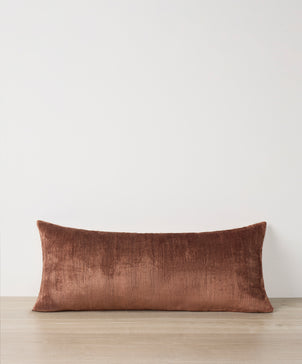 The Talik Velvet Lumbar Cushion Cover in Chestnut, handcrafted in Turkey, adds elegance as it rests on a light wooden surface against a plain white wall.