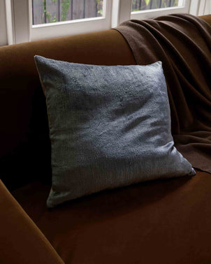 A Talik Velvet Square Cushion Cover in Mist, handcrafted in Turkey, sits on a brown sofa with a matching brown blanket, near a window where natural light softly illuminates the scene.