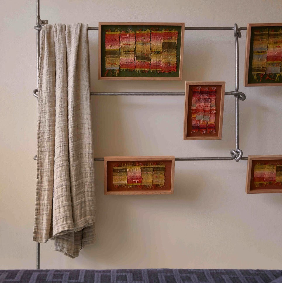 A rustic wall-mounted shelving system made of silver metal pipes and wooden frames, holding colorful rectangular art pieces and a beige textured throw blanket, against a cream wall.