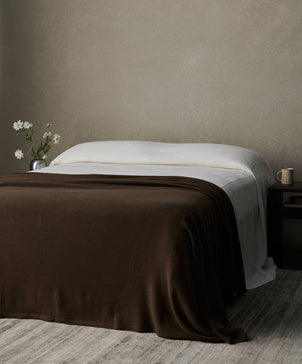 A neatly made bed with white sheets and a dark brown 100% NZ Merino Wool Blanket - Truffle sits against a plain beige wall. A nightstand holds a mug, book, and vase with white flowers. A light-colored rug lies on the floor.