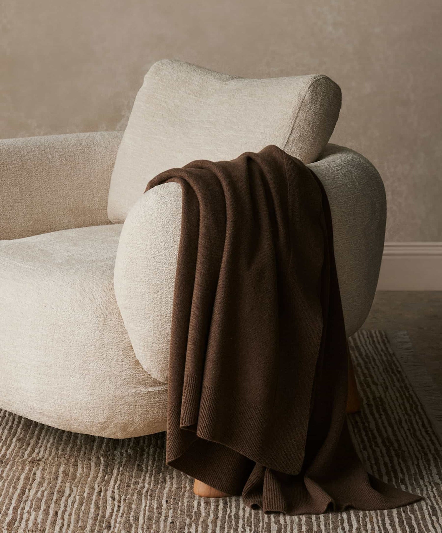 A cozy, cream-colored textured armchair with a 100% NZ Merino Wool Throw in Truffle draped over one arm, set atop a light patterned rug and positioned against a neutral wall.
