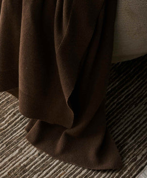 A 100% NZ Merino Wool Throw in Truffle draped over the edge of a light-colored sofa rests on a textured, striped rug in neutral tones.