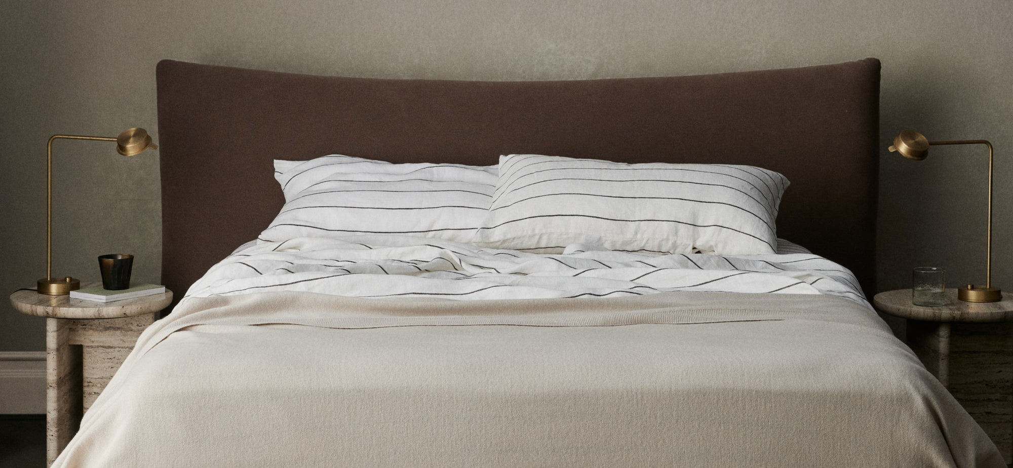 A neatly made bed with striped bedding, two pillows, a brown headboard, and matching side tables each with a gold lamp. A book, candle, and glass sit on the tables against a neutral wall.
