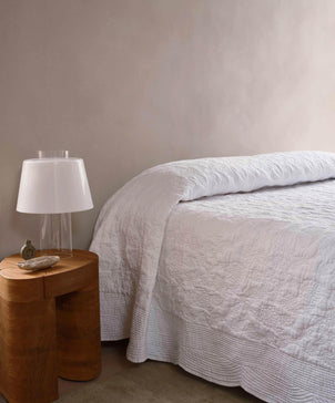 White textured blanket draped over a bed with a side table in the background.