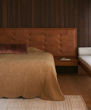 A cozy bedroom featuring the Willow Embroidered Quilt in nutmeg, a tufted leather headboard, dark wood-paneled wall, long burgundy pillow, light beige bench, and a floating nightstand with a book and eyeglasses.