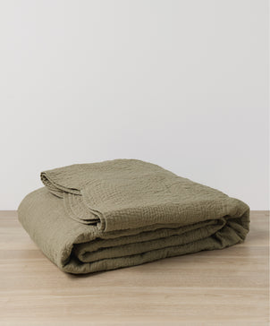 A folded Willow Embroidered Quilt - Putty with a textured, stitched pattern rests on a light wooden surface against a plain white background.