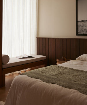 A minimalist bedroom with a bed featuring crisp white linens and the Willow Embroidered Quilt in Putty, a wooden bench by a curtained window, an open book, and natural light softly illuminating the space.