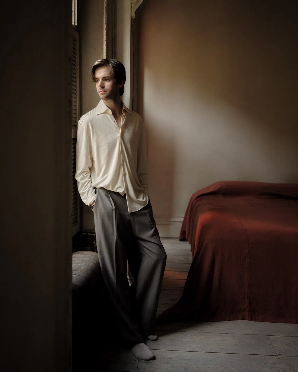 A person in a cream shirt and gray trousers stands by a window in a dimly lit room with a bed covered in a red blanket. They look outside, hands in pockets, and the atmosphere is calm and contemplative.