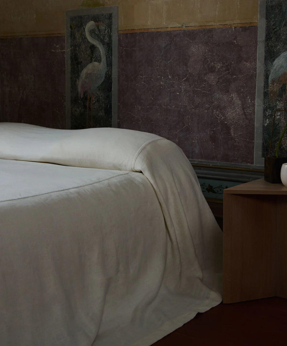 A neatly made bed with white linens next to a wooden nightstand. The wall behind has ornate panels with crane illustrations and a marbled purple section, creating an elegant, serene atmosphere.