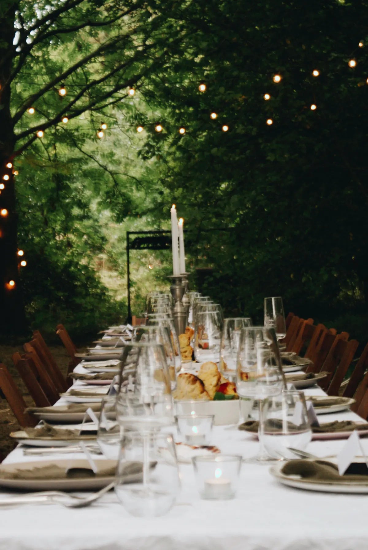 Long table set for dinner outdoors with string lights and greenery