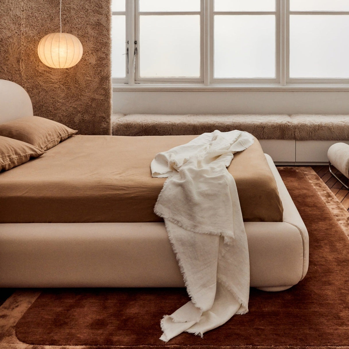 Minimalist bedroom with a beige bed, brown bedding, white throw blanket, textured wall, round hanging lamp, large windows, and a soft brown rug.