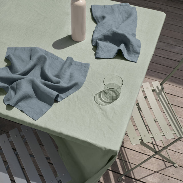 A rectangular table in an outdoor setting is dressed with a Linen Tablecloth in Sage and Linen Napkins in Bluestone.