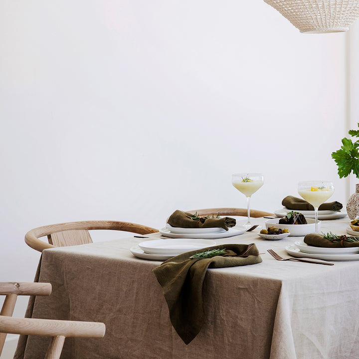 A long table setting with wooden chairs white crockery and cocktails, styled with a Natural Linen Tablecloth and Linen Table Napkins in Olive.