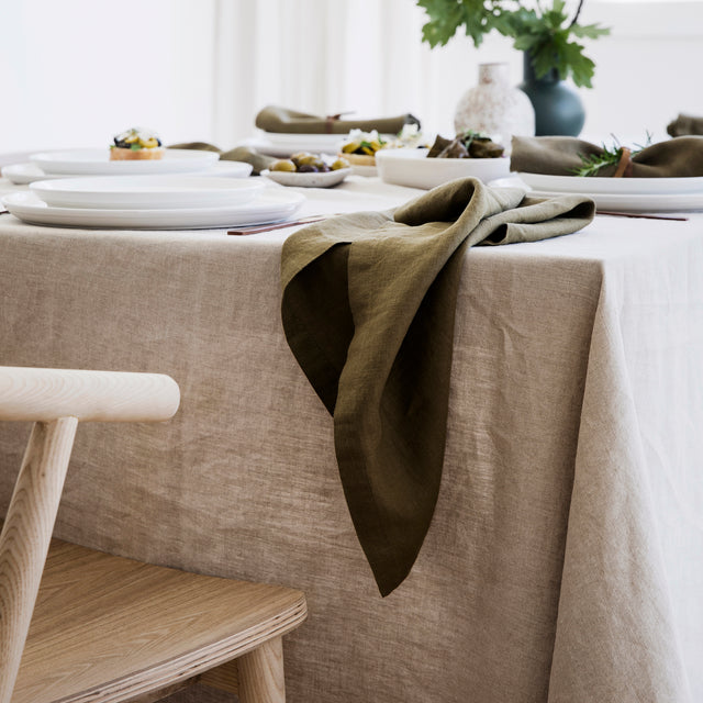 A table setting with white crockery, styled with a Natural Linen Tablecloth and Linen Table Napkins in Olive. The table is surrounded with wooden chairs.