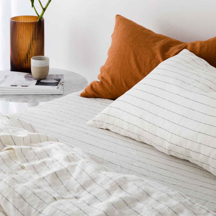 Close up of a bed dressed in Pencil Stripe bedlinen with a pillowcase in Cedar. A marble side table holds an amber glass vase and magazines.