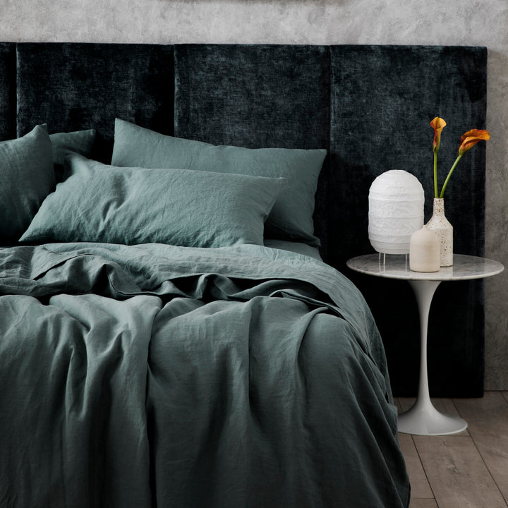 A bed dressed in Bluestone bedlinen, styled with a marble side table, a white lamp and vessels.