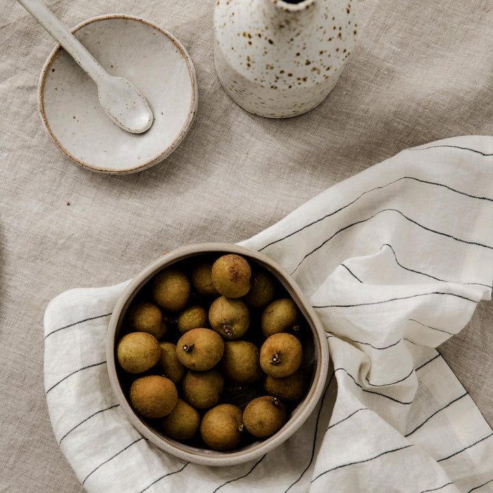 A close up table setting featuring a Natural Tablecloth, neutral textured pottery, fruit and Pencil Stripe Napkins.