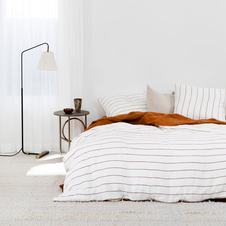 Bed styled with Cedar Stripe duvet cover set, Cedar flat sheet with border and Natural euro pillow. On the bedside table are two ceramic vases. Next to the bed is a floor lamp. Sizes: Queen, King