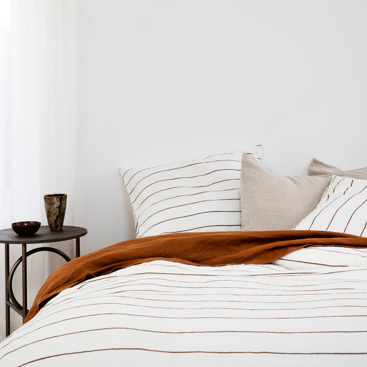 A bed dressed in Cedar Stripe, Cedar and Natural linen bedding, styled with a modern bedside table