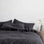A bed dressed in Slate bed linen, styled with a wooden bedside table and small grey vase. Sizes: Queen, King