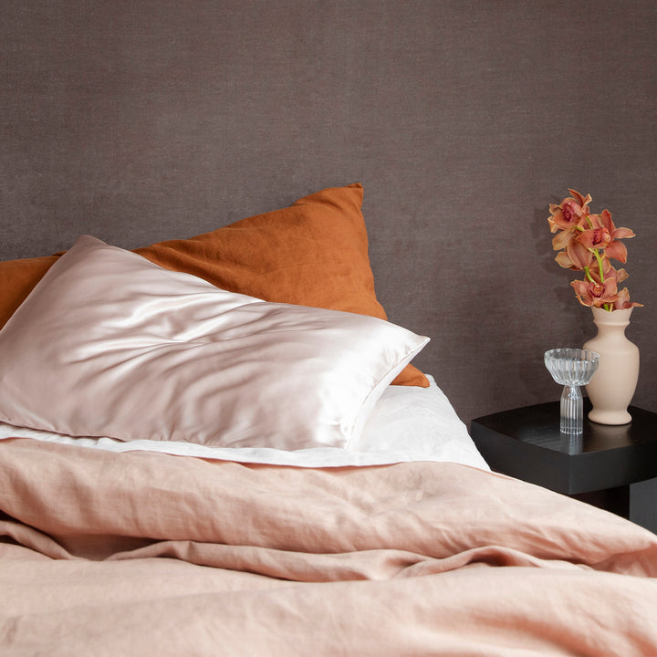 Close up of a bed dressed in Fawn and Blush bedlinen, with a pillowcase in Cedar. A black side table holds a pale pink vase with orchids.
