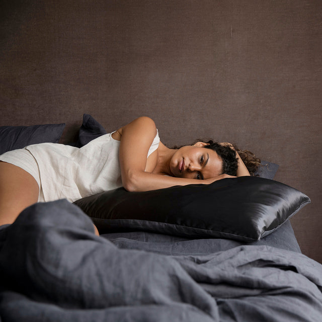 A woman relaxes on a bed dressed in Slate bedlinen, and a Silk Flip Pillowcase in Slate.