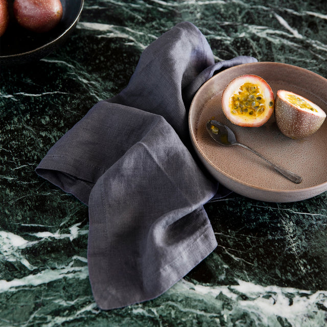 A Linen Table Napkin in Slate styled under a brown ceramic bowl with a passionfruit and silver spoon