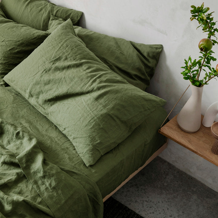 A close up on the corner of bed styled with the Linen Sheet Set with Pillowcases and Set of 2 Linen Pillowcases in Forest. Next to the bed is a floating wooden shelf styled with a white vase containing some greenery