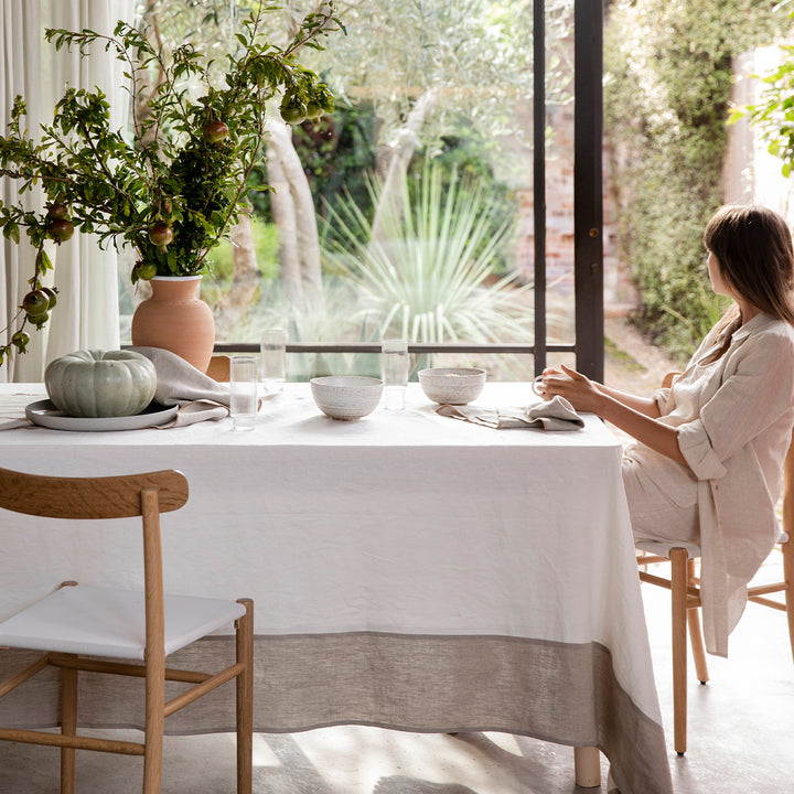 Model dressed in all beige linen sitting at a table covered in the Cara Panel Tablecloth. On top of the table are some ceramic bowls, glasses, Cara Edged Napkins in Natural, a terracotta vase of pomegranates and a large pumpkin on a gray plate/dish. The model is looking out the window at the greenery outside.