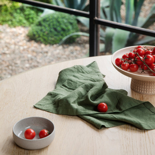 A Linen Table Napkin in Forest on a wooden table. Next to the napkin is a small dish of tomatoes and a larger stand of tomatoes.
