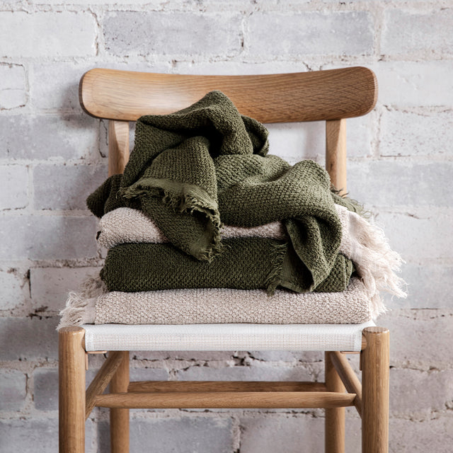 A stack of styled Pure Linen Bath Towels in Natural and Forest on a wooden chair.