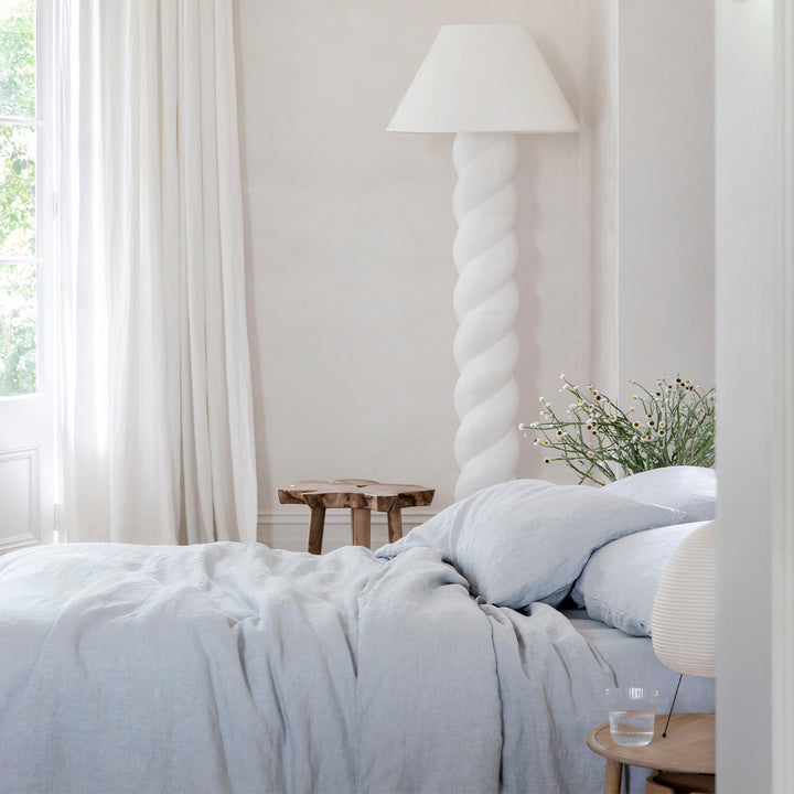 A bed dressed in Sky bed linen, styled with wooden bedside tables and white curtains. Sizes: Queen, King