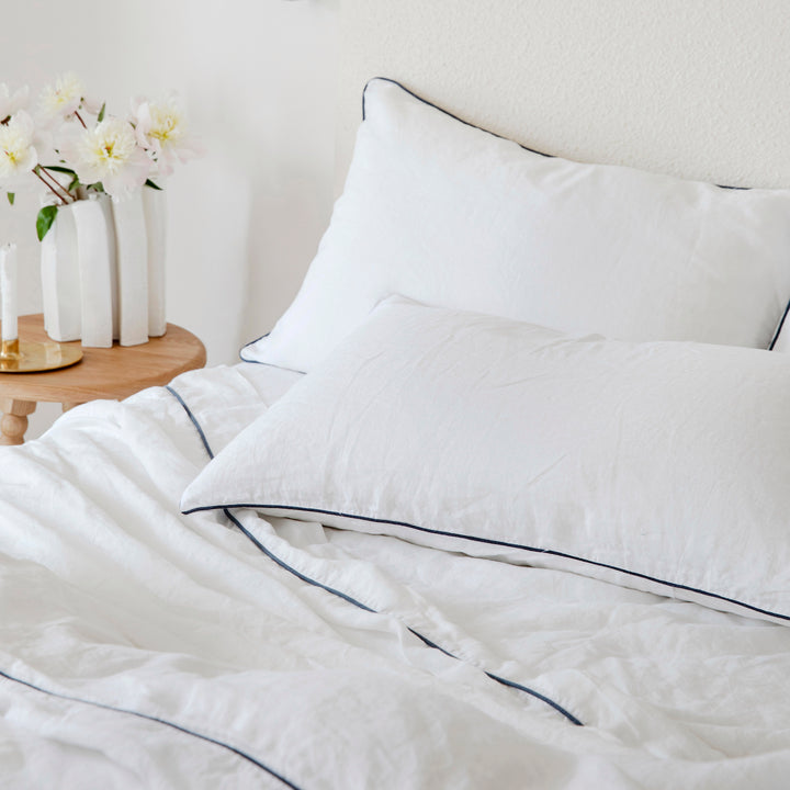 A bed dressed in White with Navy Piped bedlinen, styled with a vase of flowers on side table.