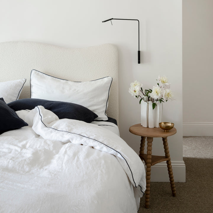 A bed dressed in White with Navy Piped bedlinen and Navy Pillowcases is styled with a vase of flowers on side table.
