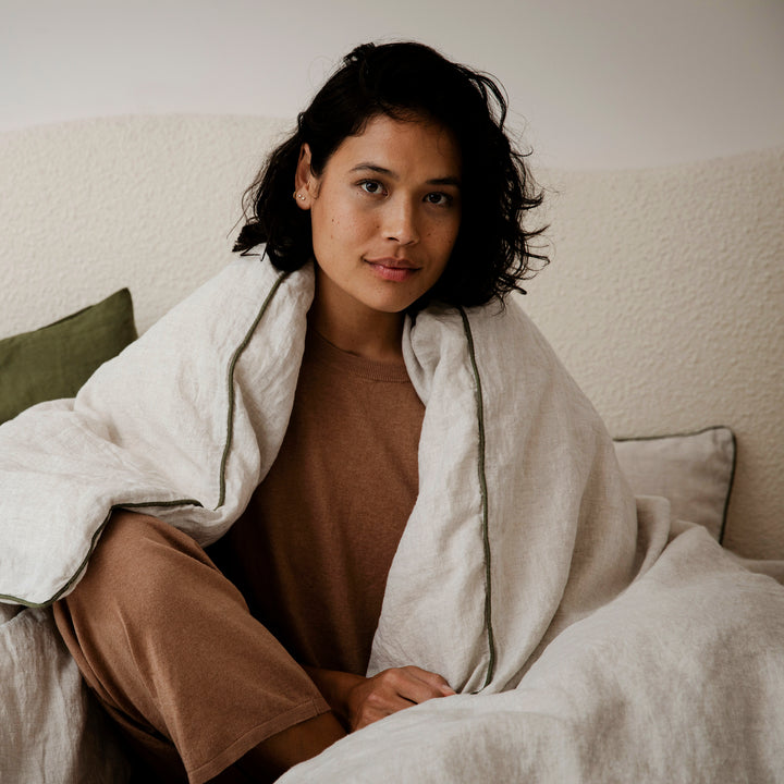 A woman is sitting on a bed dressed in Natural with Forest Piped bedlinen, and Forest Pillowcases.