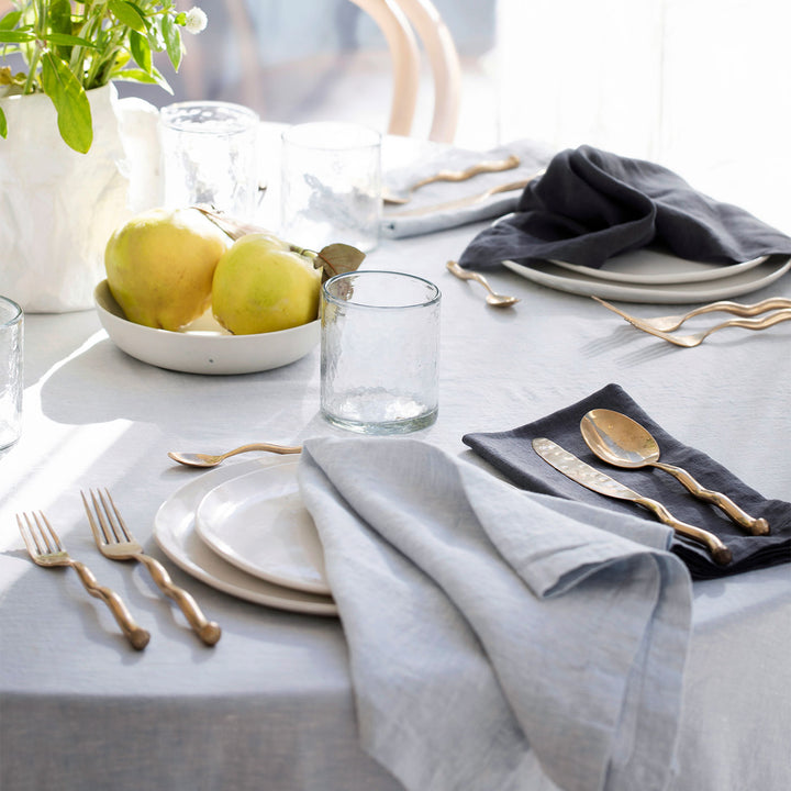 A round table is dressed in a Linen Tablecloth in Sky and Linen Napkins in Sky and Slate. The table is set with gold coloured cutlery and ceramic dinnerware